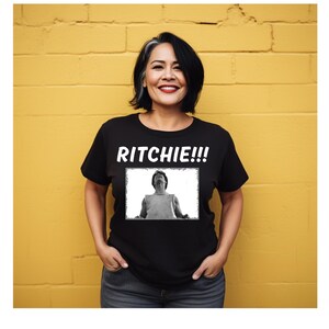 Könnte beinhalten: Schwarzes T-Shirt mit dem Wort "RITCHIE!!!" in weißen Blockbuchstaben über einem Schwarz-Weiß-Bild einer Person, die nach oben schaut. Die Person auf dem Bild trägt ein schwarzes T-Shirt und blaue Jeans. Der Hintergrund ist eine gelbe Backsteinmauer.