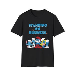 May include: Black t-shirt featuring a graphic of several Smurfs characters. The text "STANDING ON BUSINESS" is printed in blue above the characters. The Smurfs are in various poses, with some holding items.