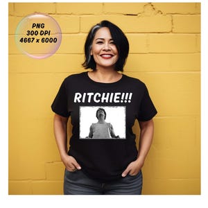 May include: A black t-shirt with the word "RITCHIE!!!" in white block letters above a black and white image of a person looking up. The background is a yellow brick wall.