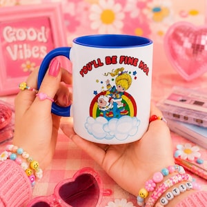 May include: White ceramic mug with a blue interior and handle, featuring a colorful illustration of a cartoon character and the text "YOU'LL BE FINE HO". The mug is held by a person with pink nail polish and multiple bracelets.