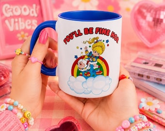 May include: White ceramic mug with a blue interior and handle, featuring a colorful illustration of a cartoon character and the text "YOU'LL BE FINE HO". The mug is held by a person with pink nail polish and multiple bracelets.