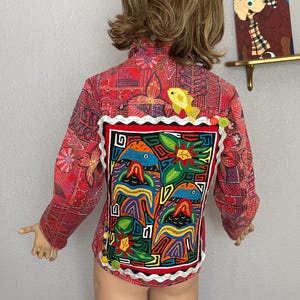 Könnte beinhalten: Rote Jacke mit farbenfrohen Stickereien. Der Rücken zeigt eine lebendige, gerahmte Applikation mit Vogel- und Blumendesigns. Die Jacke hat eine weiße Spitzenborte und ist mit bunten Knöpfen verziert. Eine gelbe Vogelapplikation befindet sich auf dem oberen Rücken.