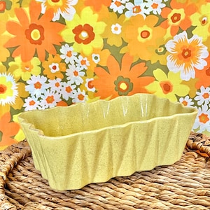 May include: A yellow ceramic planter with a wavy, ruffled edge. The planter is sitting on a woven wicker surface.