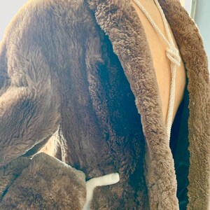 Pu&ograve; includere: Un cappotto marrone in finta pelliccia con collo a scialle e maniche lunghe. Il cappotto ha una cintura bianca e una fodera nera. Una lunga collana di perline bianche &egrave; drappeggiata sul cappotto.