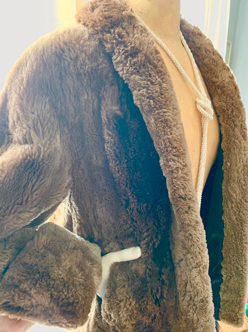Pu&ograve; includere: Un cappotto marrone in finta pelliccia con collo a scialle e maniche lunghe. Il cappotto ha una cintura bianca e una fodera nera. Una lunga collana di perline bianche &egrave; drappeggiata sul cappotto.