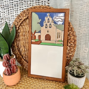 May include: A wooden framed tile with a colorful image of a church with a white background. The tile is on a woven surface with a green plant in a white pot.