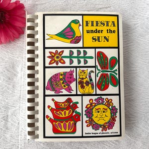 Könnte beinhalten: Ein spiralgebundenes Kochbuch mit dem Titel "Fiesta Under the Sun" mit farbenfrohen Illustrationen eines Vogels, Schweins, einer Katze und anderen festlichen Designs. Das Cover hat ein gelbes, grünes und rosa Farbschema, mit dem Text in Schwarz.