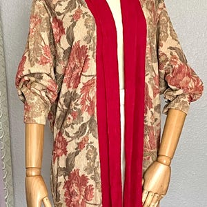 May include: A long-sleeved cardigan with a floral print in shades of brown and pink. The cardigan has a red trim along the front opening and a relaxed fit.