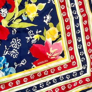 May include: A square scarf featuring a navy blue background with a floral pattern in red, yellow, and light blue. The border consists of alternating stripes of red, white, and navy blue with small white flower motifs. The name ADOLFO is printed.