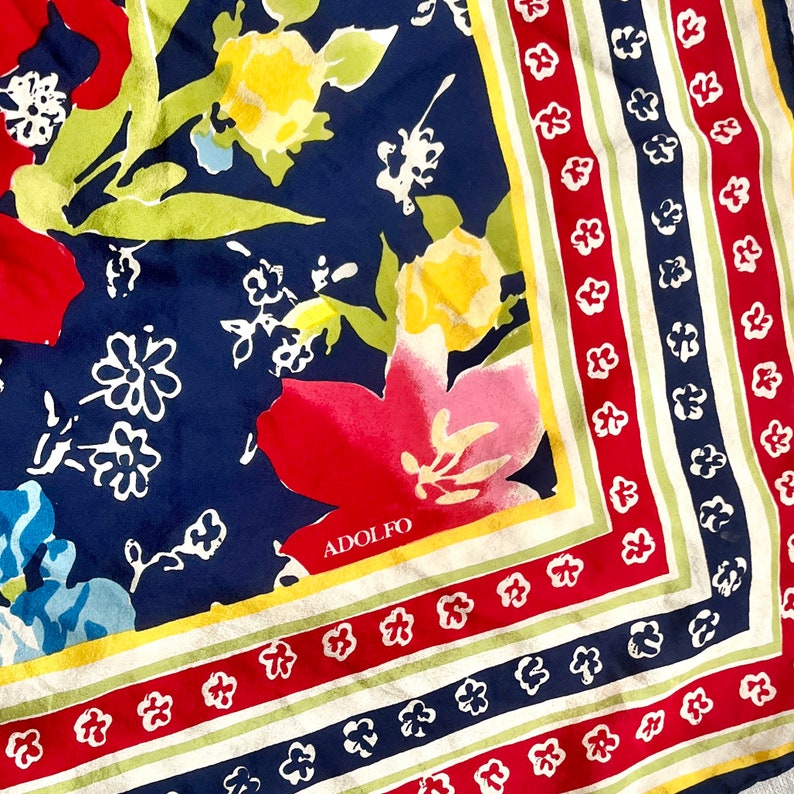 May include: A square scarf featuring a navy blue background with a floral pattern in red, yellow, and light blue. The border consists of alternating stripes of red, white, and navy blue with small white flower motifs. The name ADOLFO is printed.