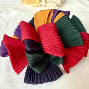May include: A multi-coloured pleated fabric flower in shades of red, purple, green, gold, and navy. The flower is made of layered, folded fabric, creating a textured, dimensional effect. The petals are arranged in a circular pattern.