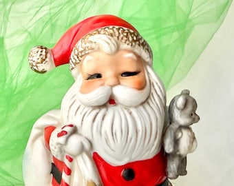 Op de afbeelding: Een keramieken kerstmanbeeldje met een rode hoed en jas, met een zak en een kleine grijze teddybeer. De kerstman heeft een witte baard en snor, met roze wangen en gesloten ogen. De achtergrond is lichtgroen.