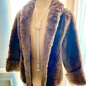 Pu&ograve; includere: Un cappotto marrone in finta pelliccia con collo a scialle e polsini risvoltati. Una lunga collana di perline bianche con un design annodato e estremit&agrave; a nappa pende dal cappotto. Il cappotto &egrave; esposto su un manichino.