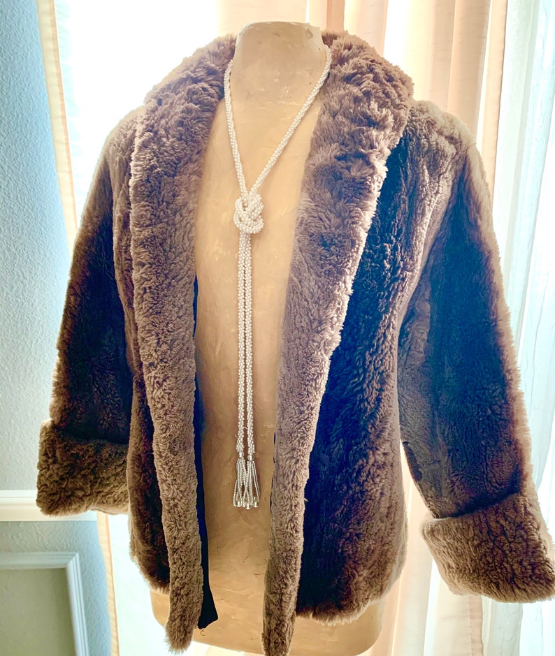 Pu&ograve; includere: Un cappotto marrone in finta pelliccia con collo a scialle e polsini risvoltati. Una lunga collana di perline bianche con un design annodato e estremit&agrave; a nappa pende dal cappotto. Il cappotto &egrave; esposto su un manichino.