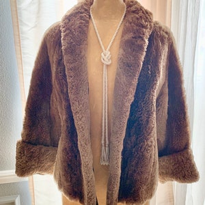 Pu&ograve; includere: Un cappotto in finta pelliccia marrone con ampio colletto e polsini ripiegati. Una lunga collana di perle bianche con un design annodato &egrave; appesa davanti al cappotto. Il cappotto &egrave; esposto su un manichino.