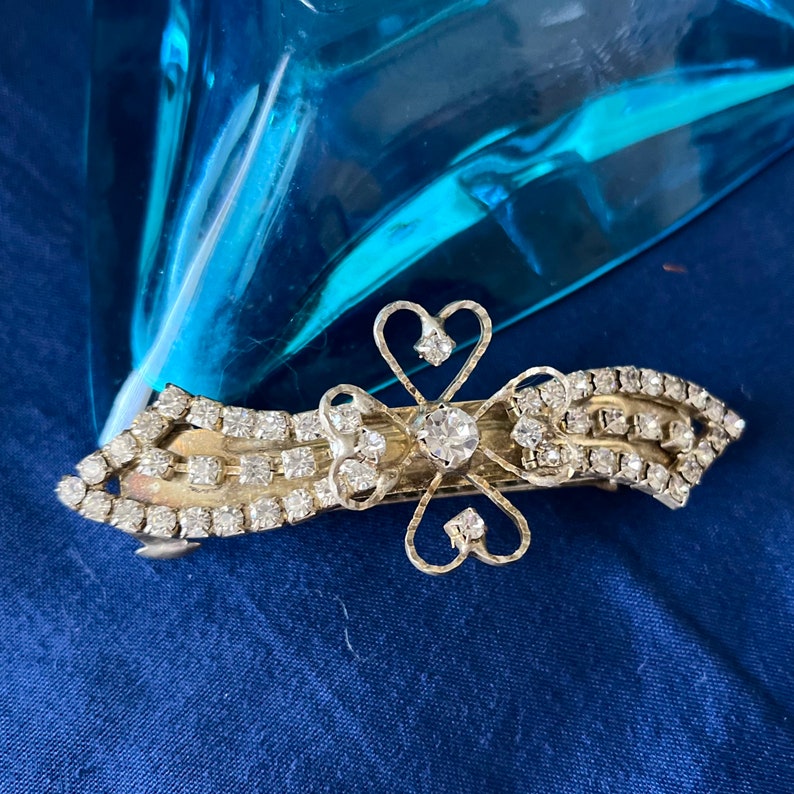May include: A silver hair clip with a decorative design of a four-leaf clover with hearts. The clip is adorned with clear rhinestones.