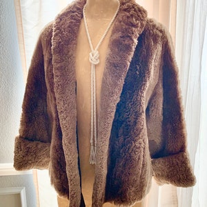 Pu&ograve; includere: Un cappotto marrone in finta pelliccia con collo a scialle e polsini risvoltati. Una lunga collana di perline bianche con un design a nodo e estremit&agrave; a nappa &egrave; appesa davanti al cappotto. Il cappotto &egrave; esposto su un manichino.