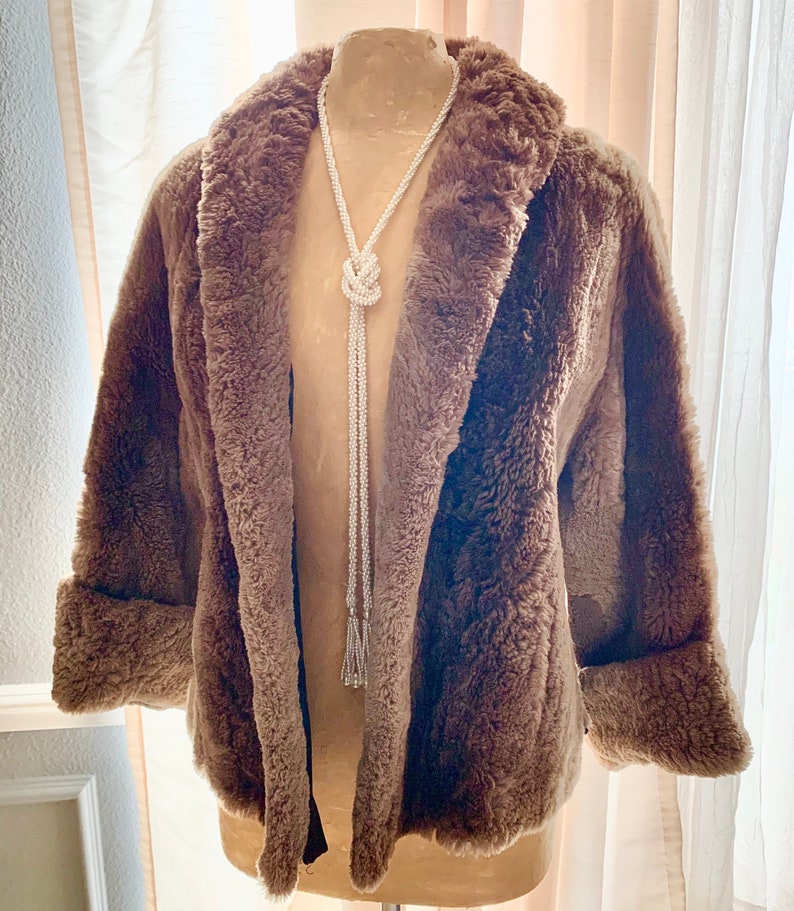 Pu&ograve; includere: Un cappotto marrone in finta pelliccia con collo a scialle e polsini risvoltati. Una lunga collana di perline bianche con un design a nodo e estremit&agrave; a nappa &egrave; appesa davanti al cappotto. Il cappotto &egrave; esposto su un manichino.