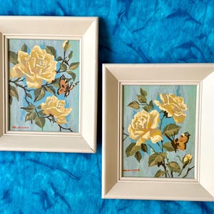 May include: Two framed paintings featuring yellow roses and butterflies against a light blue background. Each painting is encased in a simple off-white wooden frame. The artwork displays detailed floral motifs with green leaves and orange-toned butterflies.