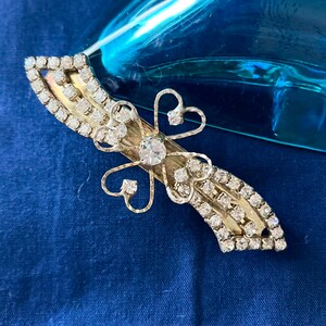 May include: A silver hair clip with a rhinestone design. The clip features a four-leaf clover shape with a large rhinestone in the center.