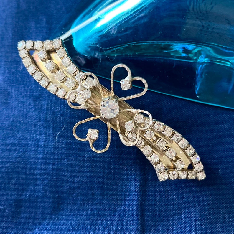 May include: A silver hair clip with a rhinestone design. The clip features a four-leaf clover shape with a large rhinestone in the center.