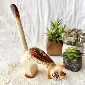 Könnte beinhalten: Handbemalte Holzkatzenfigur in Creme mit langem Schwanz. Die Katze hat rote, grüne und goldene Akzente auf dem Rücken und schwarze und rote Details im Gesicht. Die Figur wird mit kleinen Topfpflanzen präsentiert.