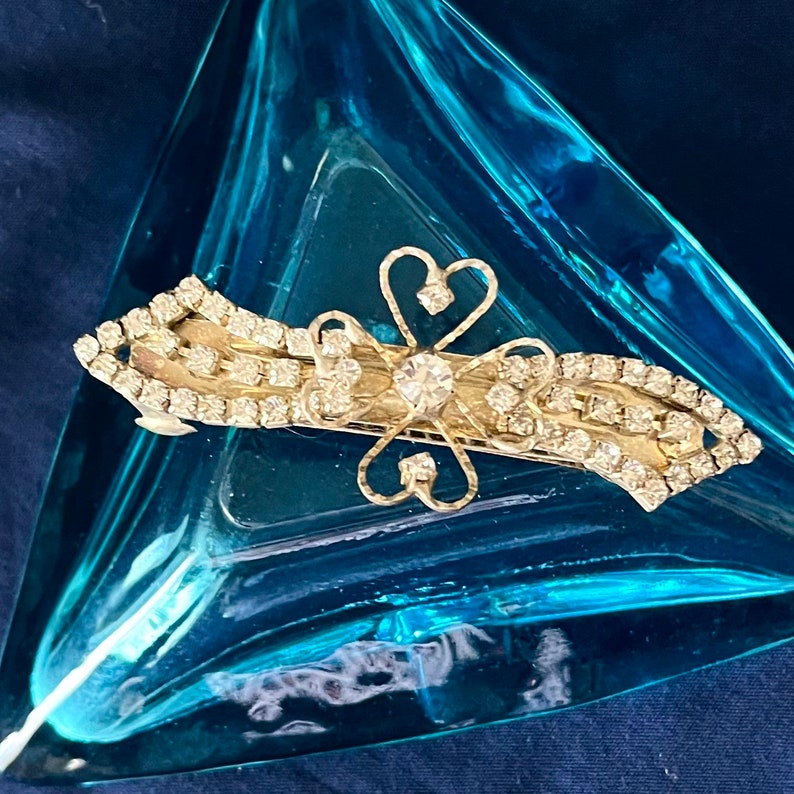 May include: A gold hair clip with a rhinestone design. The clip has a flower shape in the center with a large rhinestone in the middle. The clip is surrounded by smaller rhinestones.
