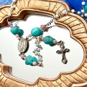 May include: A turquoise and pink beaded rosary with a silver charm and a silver crucifix. The rosary is displayed on a gold-colored mirror.