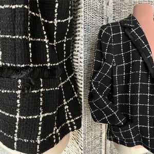 K&ouml;nnte beinhalten: Schwarze und wei&szlig;e Tweedjacke mit kariertem Muster. Die Jacke hat einen Kragen, lange &Auml;rmel und Taschen.