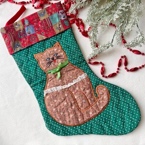 May include: A quilted Christmas stocking in green with white polka dots, featuring a brown floral cat appliqué with a green ribbon bow and white lace trim. The cuff is red with a multi-colored holiday pattern and a red fabric hanging loop.