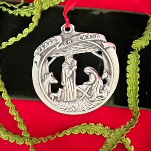 May include: A silver Christmas ornament with a scene of the nativity. The ornament is round and has a green ribbon attached to it. The ornament has the words "Peace on Earth" inscribed around the edge.