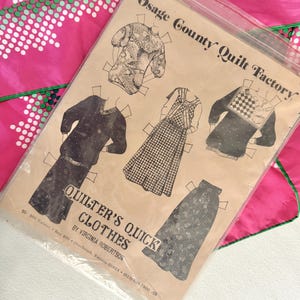 May include: A vintage sewing pattern titled "Quilter's Quick Clothes" by Virginia Robertson, featuring paper doll-style illustrations of various clothing items. The pattern is in a clear plastic sleeve, with a pink and green patterned fabric in the background.