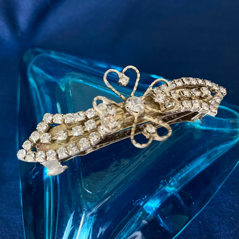 May include: A silver hair clip with a decorative butterfly design. The butterfly is made of wire and has a rhinestone in the center. The clip is adorned with rows of rhinestones.