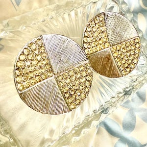 May include: A pair of silver-toned, round earrings. Each earring is divided into four sections: two sections are covered in small, sparkling yellow gemstones, and the other two have a textured, linear design. The earrings are displayed on a clear, textured surface.
