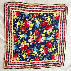 May include: A square scarf featuring a dark navy blue background with a vibrant floral pattern of red, pink, yellow, and light blue flowers and green leaves. The scarf is framed by a decorative border with red, white, and yellow stripes.