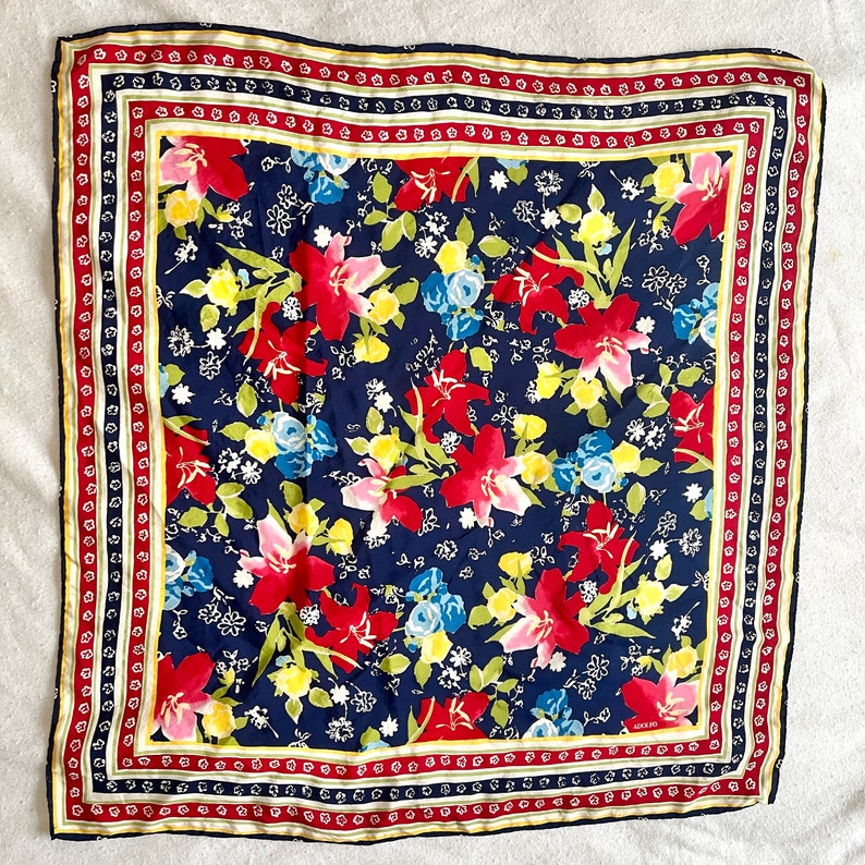 May include: A square scarf featuring a dark navy blue background with a vibrant floral pattern of red, pink, yellow, and light blue flowers and green leaves. The scarf is framed by a decorative border with red, white, and yellow stripes.