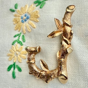 May include: A gold-coloured metal brooch in the shape of the letter "J", designed to look like a bamboo stalk with leaves. The brooch is set against a white background with embroidered yellow flowers and green leaves.