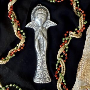 May include: A silver-toned metal angel ornament featuring a stylized figure with closed eyes, outstretched wings, and hands pressed together in a prayer position. The lower portion of the figure is decorated with embossed circular and wavy line patterns.