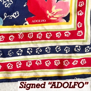 May include: A square scarf featuring a navy blue background with a large pink floral motif and the text ADOLFO. The border consists of alternating stripes in cream, light green, and red, decorated with a repeating pattern of small white flowers.
