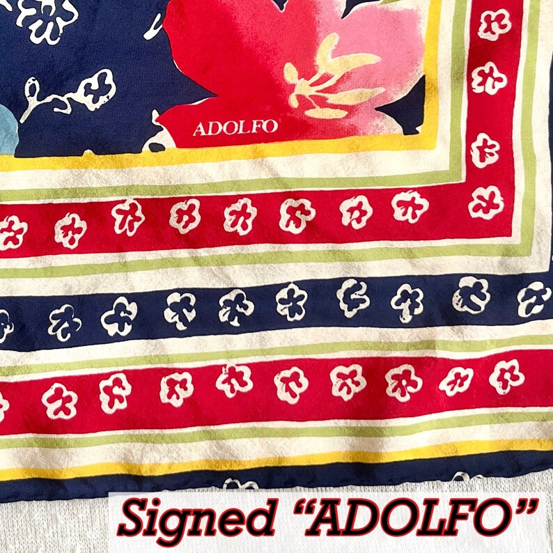 May include: A square scarf featuring a navy blue background with a large pink floral motif and the text ADOLFO. The border consists of alternating stripes in cream, light green, and red, decorated with a repeating pattern of small white flowers.