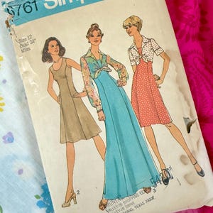 Könnte beinhalten: Vintage Simplicity Schnittmuster Nr. 761 mit drei Kleiderdesigns. Die Schnittmusterhülle zeigt Illustrationen der Kleider, mit Text, der Größe 12 und einen Brustumfang von 86 cm angibt. Die Hülle ist leicht abgenutzt, mit blau-weißem Blumenmuster im Hintergrund.