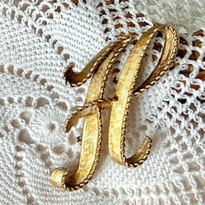 May include: A gold-toned metal brooch in the shape of the letter "H". The brooch has a textured surface and a rope-like border. The brooch is set against a white lace background.