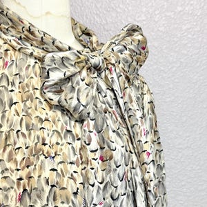 Pode incluir: Um close-up de uma blusa de seda com um padrão de penas em tons de cinza, bege e marrom. A blusa apresenta um grande laço no decote, adicionando um toque de elegância. O tecido tem um brilho suave.