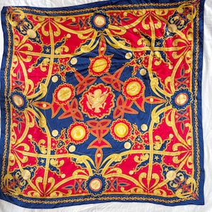 Könnte beinhalten: Ein quadratisches Seidentuch mit einem leuchtend roten und tiefblauen Hintergrund sowie komplexen goldfarbenen Ornamentmustern, darunter Lorbeerkränze, Seil-Motive und ein zentrales Adler-Emblem, umgeben von kreisförmigen Medaillons und kleinen Sternen.
