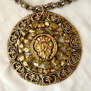 May include: A gold-toned pendant necklace with a large, ornate circular pendant. The pendant features a lion's head in the center, surrounded by intricate filigree and textured gold elements. The chain is a silver-colored, chunky link style.