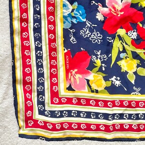 May include: A square scarf featuring a navy blue background with large red, yellow, and light green floral patterns. The border consists of alternating stripes in red, white, navy blue, and yellow with small white flower motifs. The name ADOLFO is printed in white on the navy blue section.