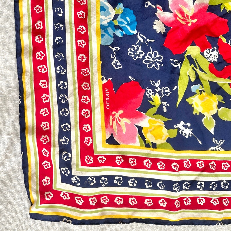 May include: A square scarf featuring a navy blue background with large red, yellow, and light green floral patterns. The border consists of alternating stripes in red, white, navy blue, and yellow with small white flower motifs. The name ADOLFO is printed in white on the navy blue section.