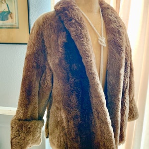 Pu&ograve; includere: Un cappotto marrone in finta pelliccia con colletto a revers e polsini ripiegati. Il cappotto &egrave; esposto su un manichino con una lunga collana di perline bianche. Il cappotto ha un aspetto morbido e strutturato e un design classico.