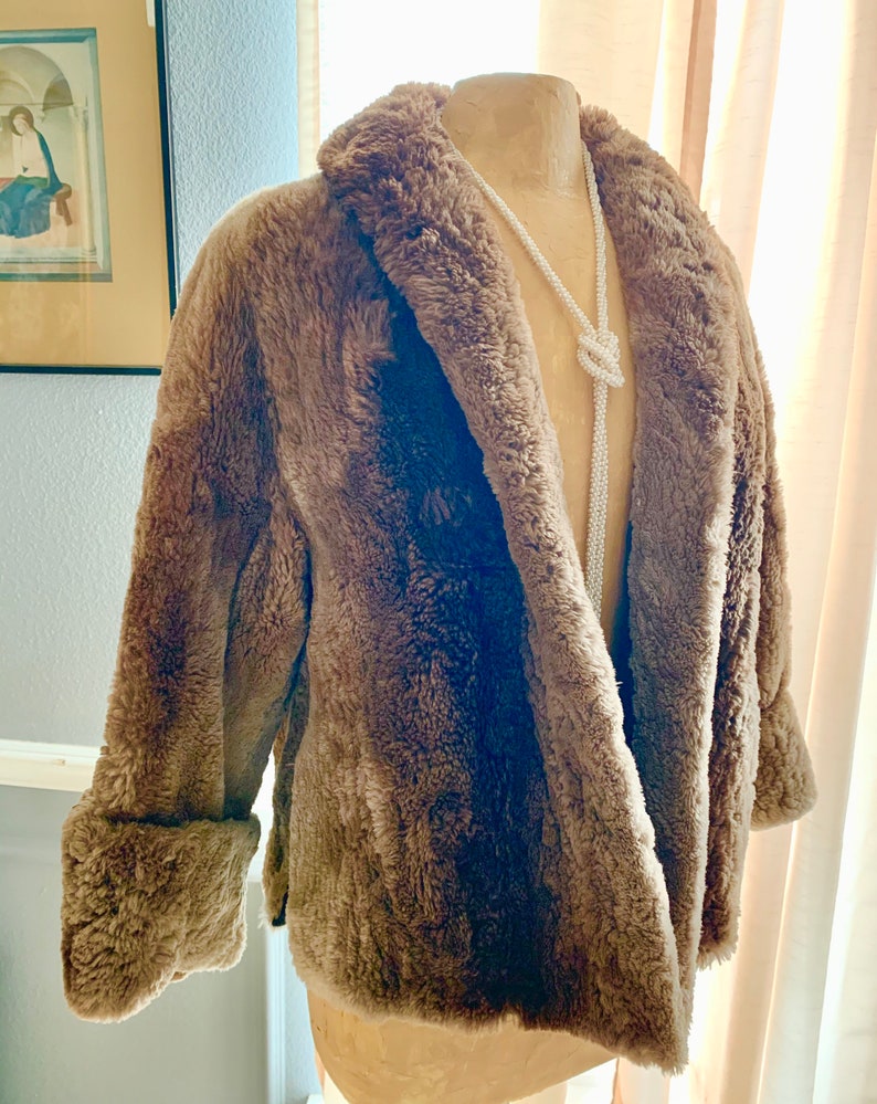 Pu&ograve; includere: Un cappotto marrone in finta pelliccia con colletto a revers e polsini ripiegati. Il cappotto &egrave; esposto su un manichino con una lunga collana di perline bianche. Il cappotto ha un aspetto morbido e strutturato e un design classico.