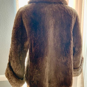 Pu&ograve; includere: Un cappotto marrone in finta pelliccia con ampio colletto e maniche lunghe. Il cappotto ha un aspetto morbido e strutturato e un design classico. Le maniche hanno un dettaglio con polsino arrotolato. Il cappotto &egrave; esposto su un manichino.